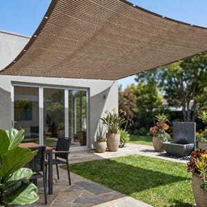 Puede incluir: Una vela de sombra rectangular de color beige con textura tejida y bordes reforzados que incluyen ojales metálicos. La tela está suspendida sobre una zona de patio, cubriendo muebles de exterior y plantas en un jardín residencial.