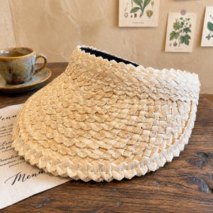 Può includere: Una visiera parasole in paglia intrecciata color beige chiaro, caratterizzata da un'ampia tesa curva e una fascia strutturata. Il materiale presenta un motivo a intreccio a cestino coerente e complesso su tutta la superficie, offrendo un aspetto testurizzato.