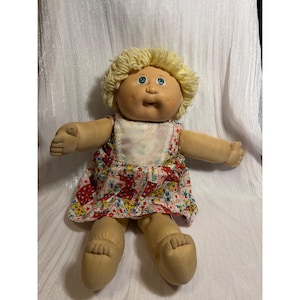 May include: A soft-bodied doll with light skin, dimpled cheeks, and blue eyes. The doll has short, curly blonde yarn hair and wears a sleeveless dress featuring a colorful patchwork pattern of red, blue, and yellow floral squares with white lace trim.