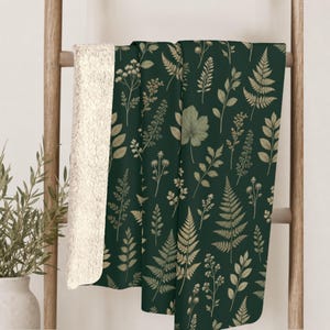 May include: A dark green throw blanket featuring a repeating pattern of light beige botanical illustrations including ferns, leaves, and wildflowers. The blanket has a thick, textured, cream-colored sherpa fleece lining visible on the folded edge.