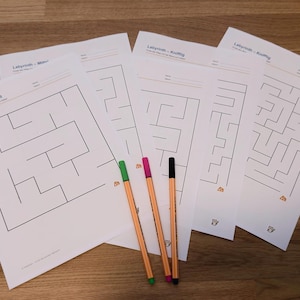 Op de afbeelding: Een set witte vellen papier met zwart gedrukte doolhofpuzzels en de tekst Labyrinth, vergezeld van vijf dunne viltstiften in groen, roze, zwart, oranje en blauw, en een kleine stapel vierkante, meerkleurige zelfklevende notitieblaadjes.