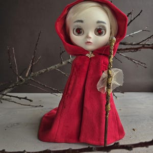 May include: A doll with a pale complexion, blonde hair, and red eyes, wearing a red hooded cloak. The cloak is fastened with a gold clasp. The doll holds a decorative staff with gold accents and a white fabric detail. The background is a neutral color with twigs.