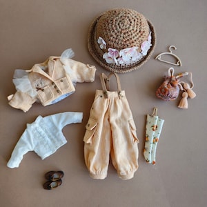 Può includere: Un set di vestiti per bambole che include una giacca beige con bordino in pizzo, una camicetta bianca, pantaloni beige con bretelle e un paio di calzini bianchi con stampa floreale.