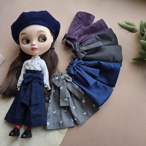 May include: A doll wearing a white shirt and blue trousers with a large bow. The doll is wearing a blue beret. There are five different pairs of blue, grey, and black trousers with bows on them.