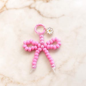 May include: A pink beaded bow keychain featuring a circular pink clasp and a small white daisy charm with a yellow center. The bow is constructed from numerous small, round, glossy pink beads strung together to form a decorative bow shape.