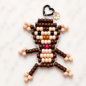 May include: A monkey-shaped keychain crafted from brown and beige plastic pony beads. The figure features a black heart-shaped lobster clasp at the top, accented with a small white and yellow daisy charm attached to the metal ring hardware.