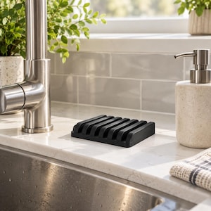 May include: A rectangular black soap dish featuring a series of parallel raised ridges designed for drainage. The soap dish has a matte finish and a solid construction, positioned on a white countertop between a metal faucet and a soap dispenser.