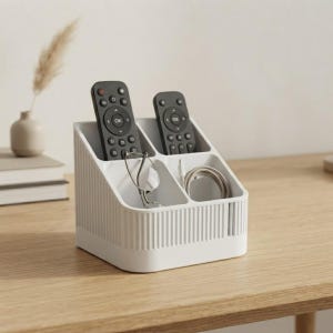 May include: A white plastic desk organiser with a ribbed exterior texture and four distinct compartments. The organiser holds two black remote controls, a pair of metal-framed spectacles, and a coiled white charging cable in its sections.