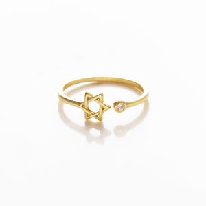 May include: A gold-colored ring featuring an open design with a Star of David and a small, round, clear gemstone. The ring has a delicate band and a polished finish, set against a white background.