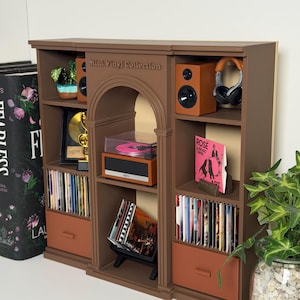 Op de afbeelding: Een bruine miniatuurkast met het opschrift Mini Vinyl Collection, voorzien van een boogvormig middengedeelte met een platenspeler, geflankeerd door zijplanken met luidsprekers, een koptelefoon, een gouden plaat en rijen miniatuur vinylhoezen.