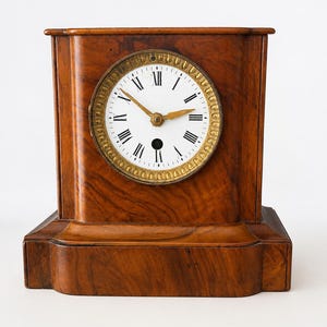 May include: A wooden mantel clock with a rectangular body and a wide base. The clock face is white with black Roman numerals and gold-colored hands, surrounded by a decorative gold-toned circular bezel. The wood features a prominent brown grain.
