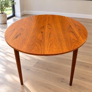 Könnte beinhalten: Ein runder Esstisch aus Holz mit einem warmen, mittelbraunen Finish und einer sichtbaren Holzmaserung. Der Tisch verfügt über eine glatte, kreisförmige Platte, die durch eine mittlere Naht geteilt ist, und steht auf vier konischen Beinen.