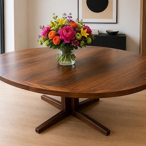 Puede incluir: Una mesa de comedor redonda de madera oscura con una superficie de grano liso y una base central en forma de cruz. Un jarrón de cristal transparente se sitúa en el centro de la mesa, lleno de un vibrante ramo de peonías rosas, rosas naranjas y lirios amarillos.