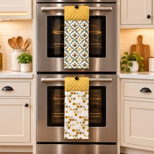 May include: Two hanging kitchen towels with yellow quilted tops and black button closures. The top towel features a pattern of bees and green leaves, while the bottom towel displays a yellow and white honeycomb pattern with small flying bees.