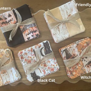 May include: Five sets of square fabric coasters tied with tan twine. The sets feature printed patterns labelled as Jack O Lantern, Friendly Ghost, Spooky Forrest, Black Cat, and Witchy Fall, each displaying seasonal motifs in orange, black, and white.
