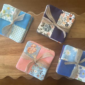 May include: Four stacks of square fabric coasters, each tied with a natural jute twine bow. The coasters feature various patterns including floral prints, solid colors, and gingham checks in shades of blue, pink, orange, and dark purple.