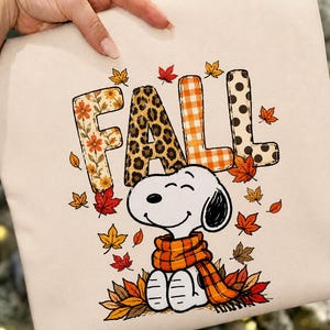 Op de afbeelding: Een beige stoffen item met het woord FALL in grote letters met diverse patronen, waaronder bloemen, luipaardprint, oranje ruitjes en zwarte stippen. Onder de tekst staat een getekende hond met een oranje sjaal tussen herfstbladeren.