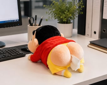 May include: A plush toy character featuring a black-haired head, a red long-sleeved shirt, and yellow shorts. The character is in a prone position with a white tissue emerging from the back of the shorts, serving as a functional tissue dispenser.