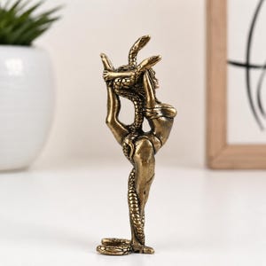 May include: A bronze-coloured figurine depicting a figure in a backbend pose with a snake coiled around the body. The figure has arms raised overhead, holding the snake, which extends down the back and legs. The sculpture has a metallic finish.