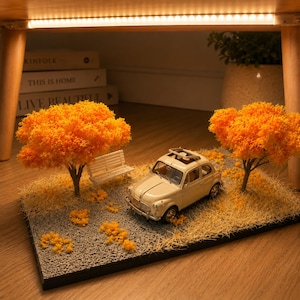 May include: A miniature cream-colored car model with a sunroof parked on a gray gravel base. The scene includes two small trees with bright orange foliage, a white bench, and scattered orange particles representing fallen leaves on the ground.