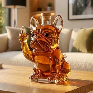 May include: A clear glass decanter shaped like a French bulldog with amber-coloured liquid inside. The dog figure has one front paw raised with the middle finger extended. The decanter features a cork stopper at the top of the head and a square base.