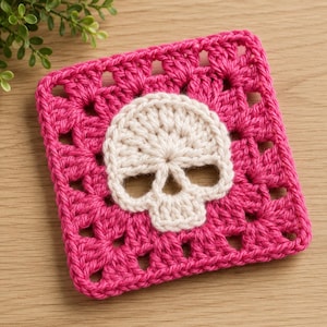 Könnte beinhalten: Ein quadratisches Häkelquadrat im Granny-Square-Stil mit einem leuchtend pinken Rand und einem zentralen, cremeweißen Totenkopfmotiv. Das Stück ist in einem klassischen Lochmuster gefertigt und erzeugt ein strukturiertes, geometrisches Design.