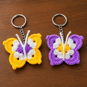 Puede incluir: Dos llaveros de mariposa tejidos a ganchillo con anillas y cadenas de metal. La mariposa izquierda tiene alas amarillas con detalles blancos y morados. La mariposa derecha tiene alas moradas con detalles blancos y amarillos. Antenas blancas.