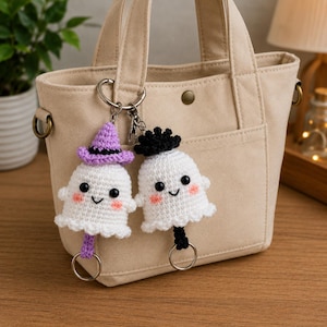 May include: Two crocheted white ghost keychains attached to a beige canvas tote bag. One ghost wears a purple witch hat, and the other has a black top hat. Both ghosts feature black bead eyes, pink stitched cheeks, and silver metal key rings.
