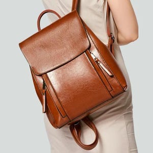 Peut inclure: Un sac à dos marron de taille moyenne avec une finition lisse et polie. Le sac présente un grand rabat supérieur avec une fermeture sécurisée, deux poches zippées verticales sur le panneau avant, une poignée supérieure et des bretelles réglables.