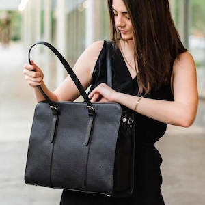 May include: A black rectangular tote bag with a pebbled texture, featuring two thin shoulder straps attached with gold-toned metal buckles. The bag has a structured silhouette with vertical stitched detailing on the front panel and side hardware.