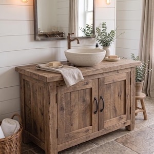Può includere: Un mobile da bagno in legno rustico con due ante e maniglie nere, sormontato da un lavabo a bacinella rotondo in pietra testurizzata color bianco sporco, un rubinetto in bronzo, un asciugamano beige e una saponetta su pannelli bianchi.