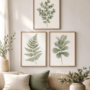 Può includere: Tre stampe artistiche verticali incorniciate con illustrazioni ad acquerello di foglie botaniche verdi, tra cui un ramo di eucalipto, una fronda di felce e un ramo di fico a foglia di violino, in cornici di legno chiaro su una parete neutra.