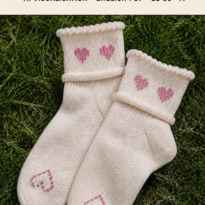 May include: A pair of off-white knit socks featuring pink heart motifs on the cuffs and toes, with a rolled edge. The image includes the text: DIGITAL KNITTING PATTERN, AI VISUALISATION, ENGLISH PDF, EU 35-41.
