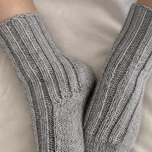 May include: A pair of light gray knitted socks with a vertical ribbed pattern. The image includes the text: DIGITAL KNITTING PATTERN, AI VISUALIZATION, ENGLISH PDF, 6 SIZES. The socks feature a textured knit construction and a reinforced heel.