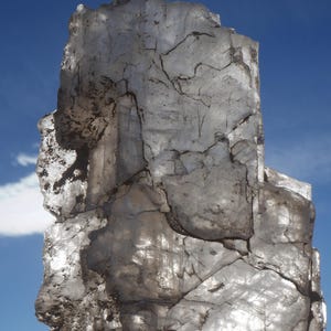 May include: A large, translucent, crystalline mineral specimen with jagged edges and internal fractures. The stone is clear with white and grey inclusions, displaying a blocky, irregular shape that allows light to pass through its surface.