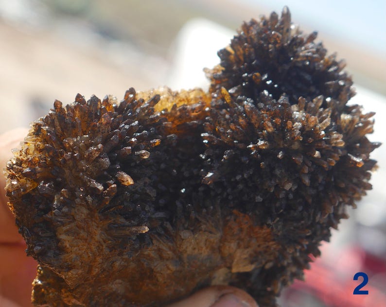May include: A cluster of dark brown, translucent, needle-like crystals forming two distinct rounded mounds on a textured, amber-coloured base. The mineral specimen features numerous sharp, pointed terminations that catch the light, creating a deep, multi-faceted appearance across the entire surface of the formation.