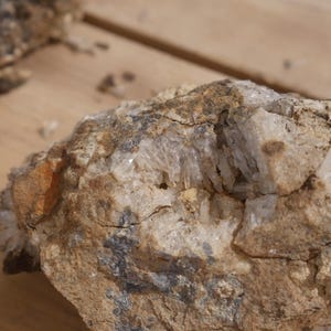 May include: A rough, earth-toned rock specimen featuring a central cavity filled with clusters of translucent, needle-like quartz crystals. The exterior of the stone displays a textured, sandy-brown surface with patches of dark grey mineral deposits.