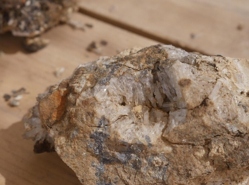 May include: A rough, earth-toned rock specimen featuring a central cavity filled with clusters of translucent, needle-like quartz crystals. The exterior of the stone displays a textured, sandy-brown surface with patches of dark grey mineral deposits.