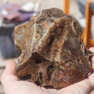 May include: A rough, irregular rock specimen featuring a deep brown and reddish-brown colour palette with crystalline, sparkling mineral deposits embedded within its crevices and along the textured, jagged surface of the stone formation.