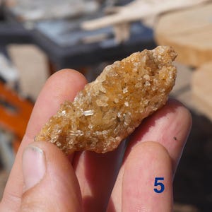 May include: A raw, elongated cluster of golden-brown crystals with a textured, multifaceted surface. The mineral specimen features numerous small, sharp, translucent points that catch the light, and it is marked with the blue number 5.