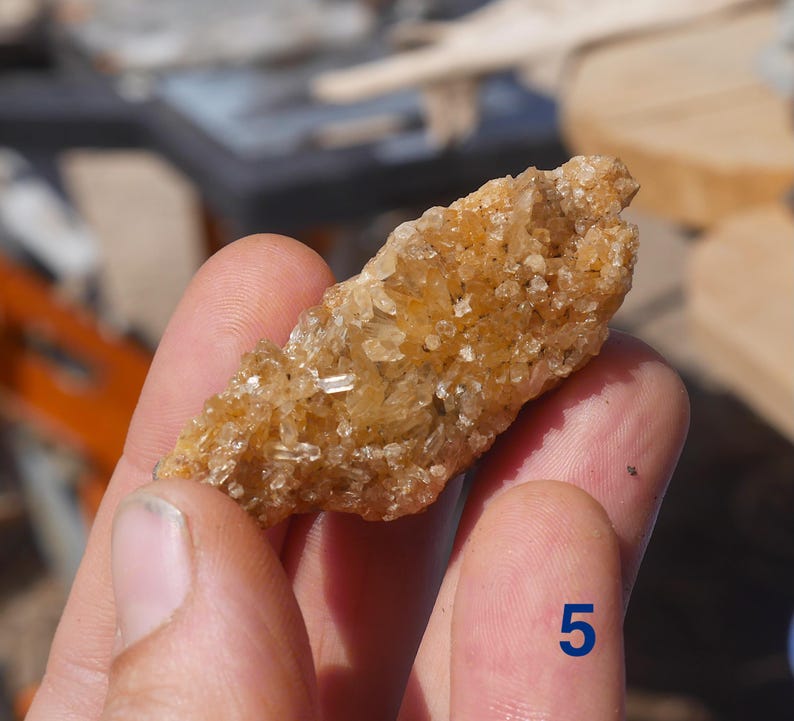 May include: A raw, elongated cluster of golden-brown crystals with a textured, multifaceted surface. The mineral specimen features numerous small, sharp, translucent points that catch the light, and it is marked with the blue number 5.