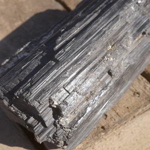 May include: A raw, black tourmaline crystal specimen featuring a deeply striated, columnar structure with a natural, jagged texture. The mineral displays a dark, opaque appearance with vertical grooves running along its length and rough edges.
