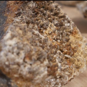 May include: A raw mineral specimen featuring a cluster of small, translucent brown and clear crystals embedded in a textured, tan-coloured matrix. The rock formation displays a jagged, uneven surface with varying shades of earthy brown and dark grey.