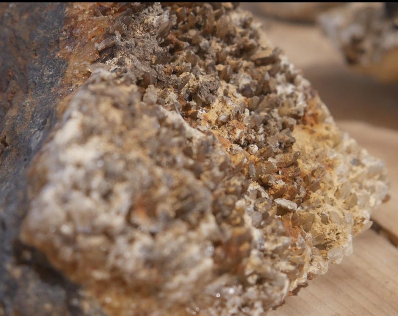 May include: A raw mineral specimen featuring a cluster of small, translucent brown and clear crystals embedded in a textured, tan-coloured matrix. The rock formation displays a jagged, uneven surface with varying shades of earthy brown and dark grey.