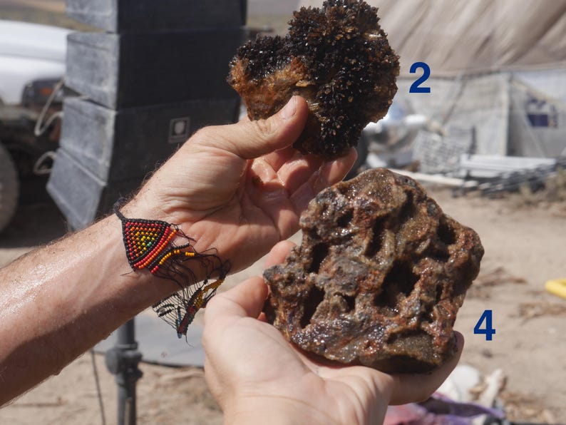 May include: Two distinct mineral specimens are held in view. Specimen 2 is a dark, crystalline cluster with a rough texture. Specimen 4 is a porous, brownish-red rock with an irregular, cavernous surface. A beaded bracelet with a rainbow pattern is visible.