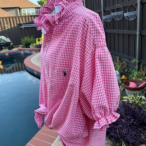 May include: A pink and white gingham button-down shirt featuring a high ruffled collar, short puff sleeves with ruffled cuffs, and a small black embroidered logo on the chest pocket. The shirt is displayed on a mannequin and has a tie-front hem.