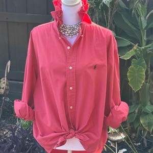 May include: A long-sleeved, button-down shirt in a solid coral-red color featuring a ruffled collar, a front button placket, and a small black embroidered logo on the left chest. The shirt is tied at the waist and styled with a multi-strand pearl necklace.