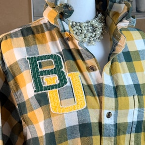 May include: A long-sleeve button-down shirt featuring a yellow, green, and white plaid pattern. The shirt has a ruffled collar and a large, stitched applique on the chest displaying the letters BU in green and yellow patterned fabric.