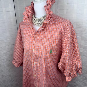 May include: An orange and white gingham button-down shirt featuring a high ruffled collar, elbow-length sleeves with ruffled cuffs, and a small green embroidered logo on the chest. The shirt is displayed on a mannequin with a pearl necklace.