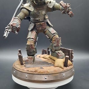 Könnte beinhalten: Eine verwitterte olivgrüne gepanzerte Figur mit rundem Helm, einem leuchtenden kreisförmigen Licht und einem Kreuzemblem auf der Brust. Die Figur hält eine mechanische Waffe und steht auf einem Sockel mit Sandsäcken, Holzplanken und Metallschrott.